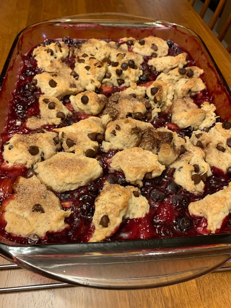 A transparent oblong pan filled with blueberries, raspberries, strawberries, and chocolate chip biscuits on a wire rack over a wooden table.