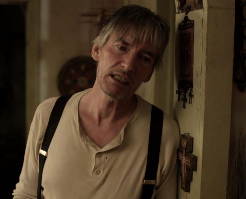 Billy Drago as the villain 'Preacher' in Children of the Corn: Genesis. He wears a stained vest with suspenders & leans against a wall with assorted religous iconography on it. There is definitely something 'off' about him, he seems awfully secretive & creepy.