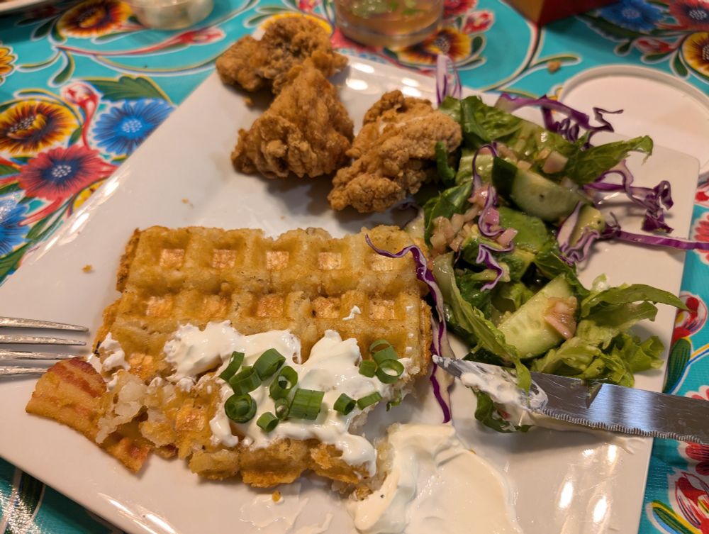 A square plate with a tater tot waffle topped with sour cream and green onion, a mixed green salad, and fried chicken. The plate is on a floral turquoise tablecloth 
