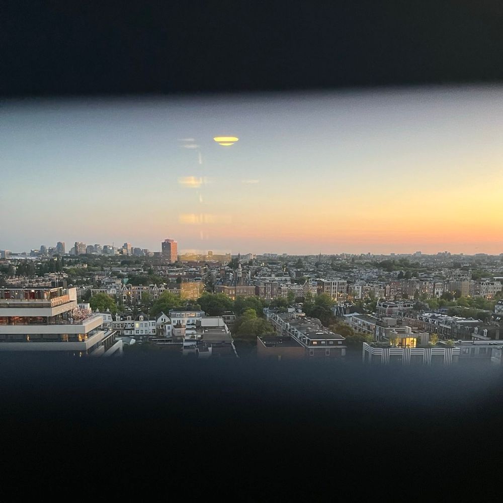 A wide sunset view over the cityscape of Amsterdam, seen from a high vantage point, with buildings and tree-lined streets stretching toward the horizon under a soft pastel sky.