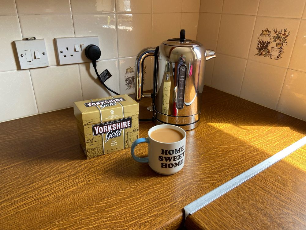 A kitchen counter top with a metallic silver electric kettle, a box of “Yorkshire Gold” tea, and a mug with tea visible in it with the words “Home Sweet Home” upon the side.