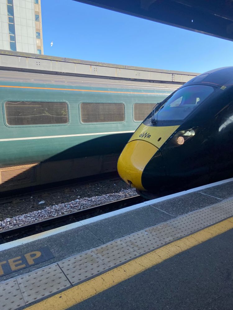 A view from a train station platform, overlooking the tracks. The very front of a green and yellow train is visible forming a curved rounded shape.