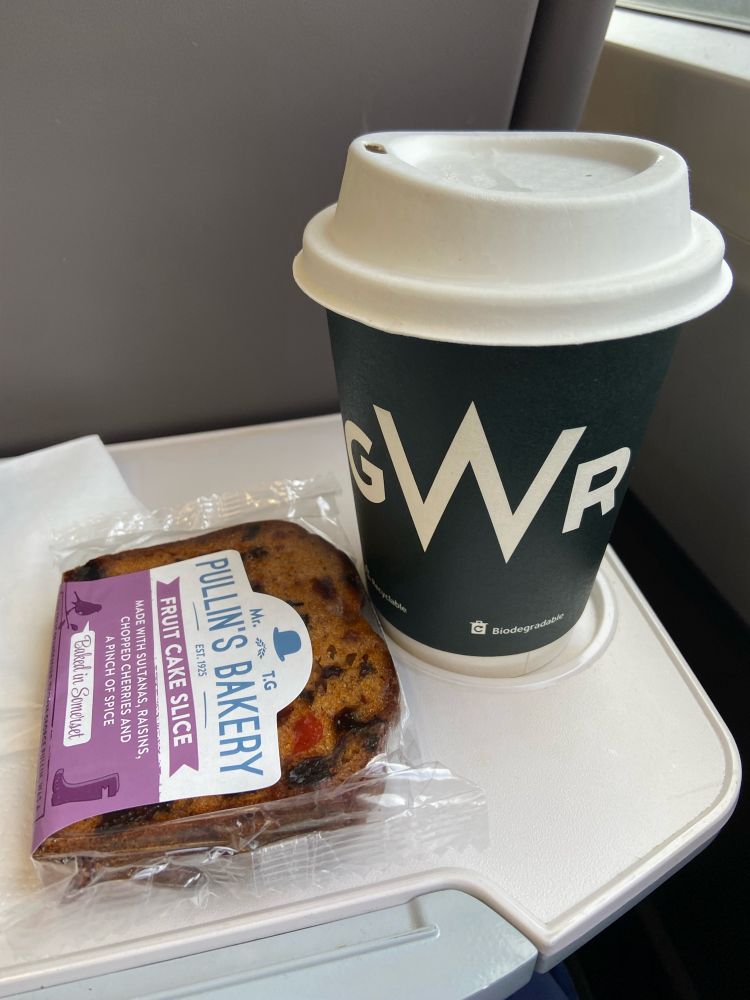 An airline style tray on the back of a train seat with a coffee mug with a large “GWR” logo on it (standing for Great Western Railway”) along with a slice of cake.