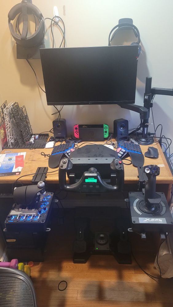 Compter desk with a monitor on a arm, an Oculus Rift S hanging on the wall, a flight yolk, flight stick, and throttle clamped to the desk and rudder peddles on the floor under the desk 