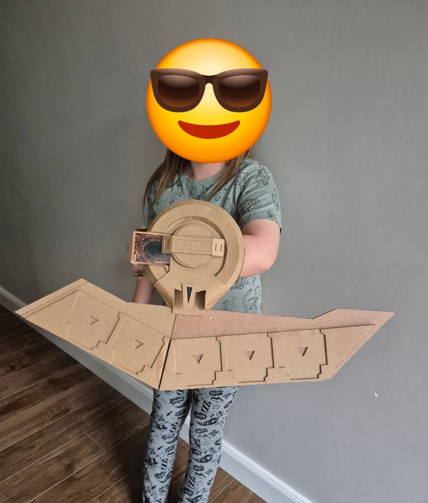 ein Kind steht vor einer grauen Wand und präsentiert an seinem linken Arm eine große Duell Disk aus Pappe. Die Duell Disk ist ein Requisit zum Spielen mit Yu-Gi-Oh Karten.