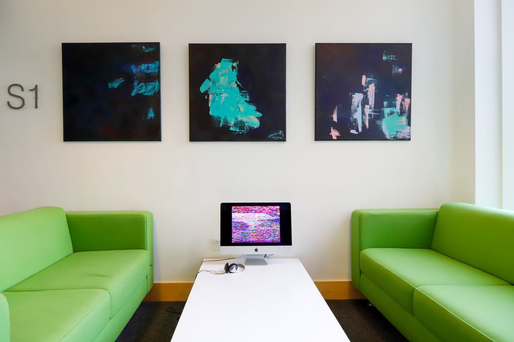 Three abstract paintings in black and turquoise on a wall and a screen on a low table showing video work.