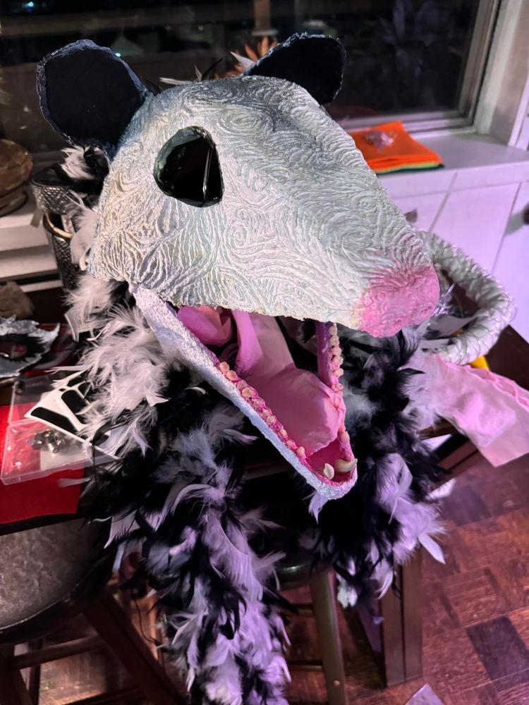 A papier-mâché stylized opossum mask laying on a table surrounded by a ratty feathered black and white boa, its jaw open as it screams into the void