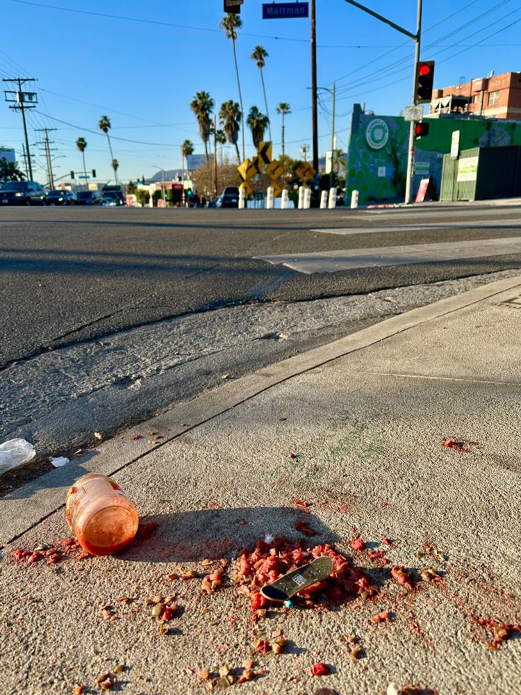 A small toy skateboard with blue wheels sits in the contents of a jar of salsa in the ramp cut of a sidewalk on a corner in Los Angeles. The jar, mostly empty, lays off to the side. The sun is shining. The events leading up to this moment are a mystery.