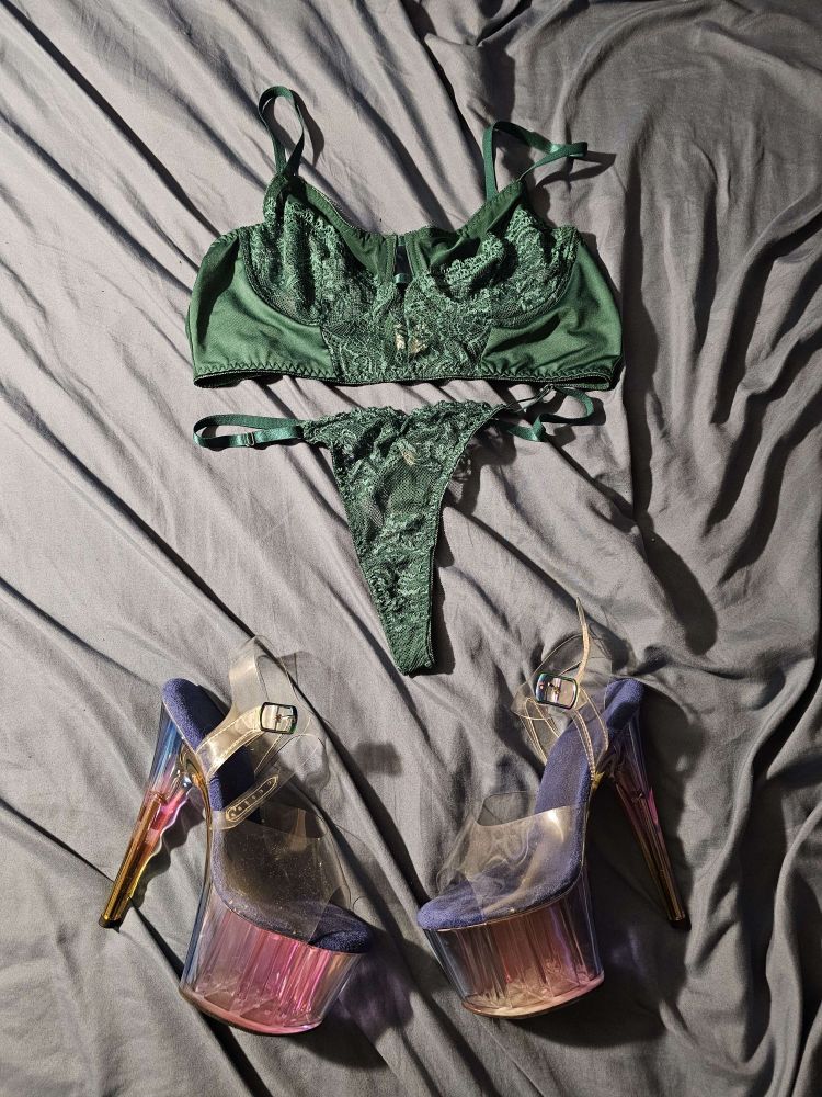 A green lacy bra and panty set and a pair of 7 inch heels are laying out on a bed with  light blue sheets.