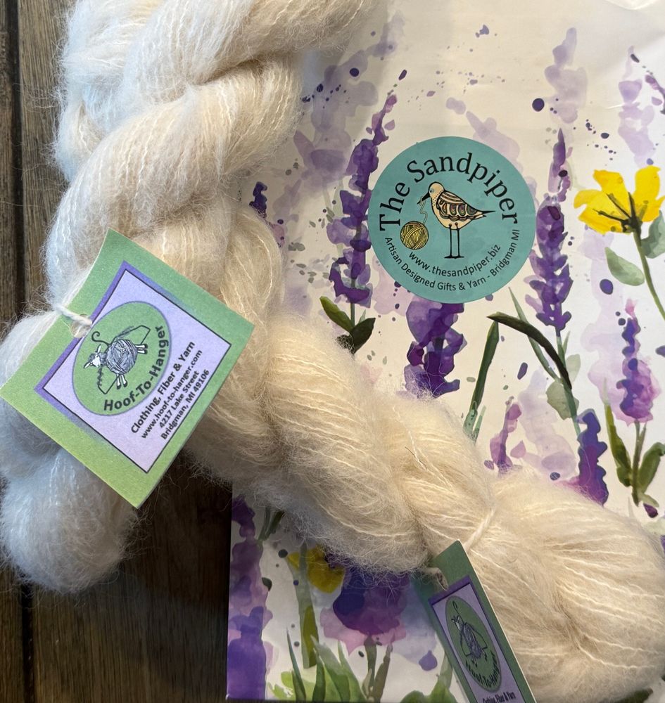2 small hanks of lace weight natural white alpaca/silk yarn from Hoof-to-Hanger shown on top of a  lavender floral shopping bag. Purchased at The Sandpiper, a gift and yarn store in Bridgman, Michigan.