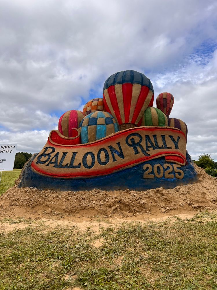 Sand sculpture of a bunch of hot air balloons rising behind a banner that says Balloon Rally 2025z the sand is colored making the balloons and banner vibrant 