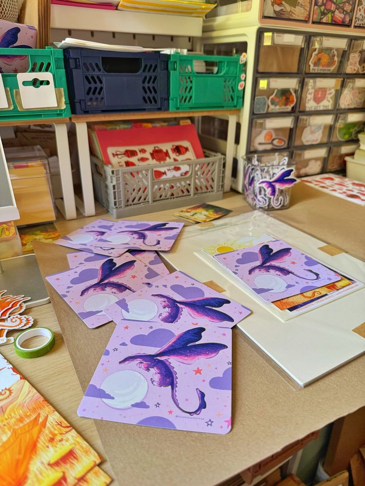 Photo shows a desk space where there is an order waiting to be wrapped in brown paper and multiple cards spread out that have a purple dragon on the front. There’s also a pot of stickers with a purple dragon sticker in and in the background are various crates and draws for organising.