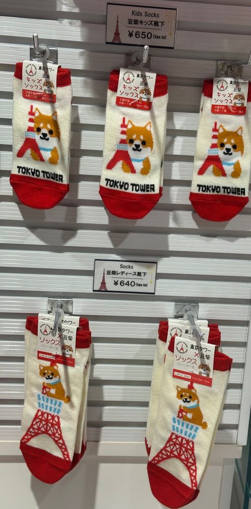 A display of socks for sale at the Tokyo Tower with a sign that says “Kids Socks” showing socks with a dog next to the tower printed on them & a sign that just says “Socks” with a dog humping the tower printed on them 