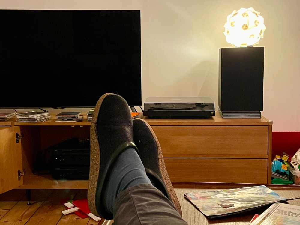 Feet in blue socks and. black felt slippers with cork soles in front of stereo, record player and TV media ensemble on wooden shelf. Stacks of CDs in front of TV. Duplo on the wooden floor, Asterix comic on the table under said feet. A cheap, glowing plastic ball lamp is laying on a black speaker.