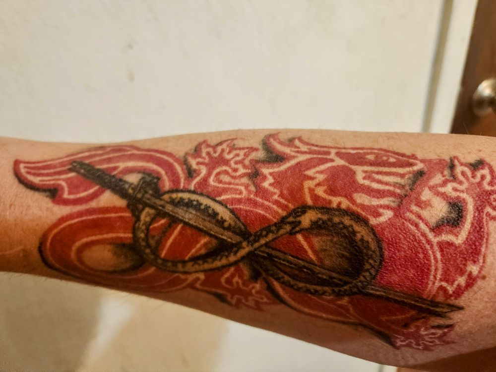 A right forearm tattoo of a red Chinese like dragon, which is a replica of the wheel.of time Rand chapter icon. On top of the dragon is a heron marked blade, that is katana like, surrounded by an oroborus in the infinity symbol. 