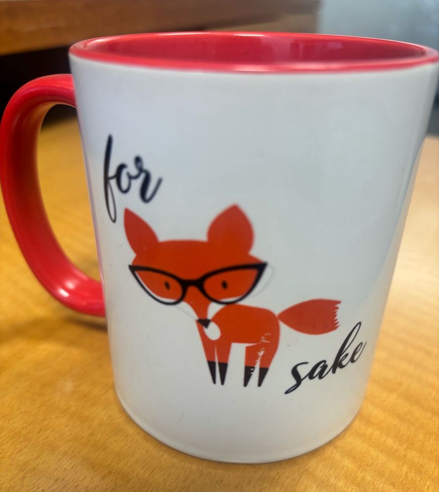 White mug orange handle. Says "for 🦊sake" with the fox as a picture of the animal with orange fur and black glasses 