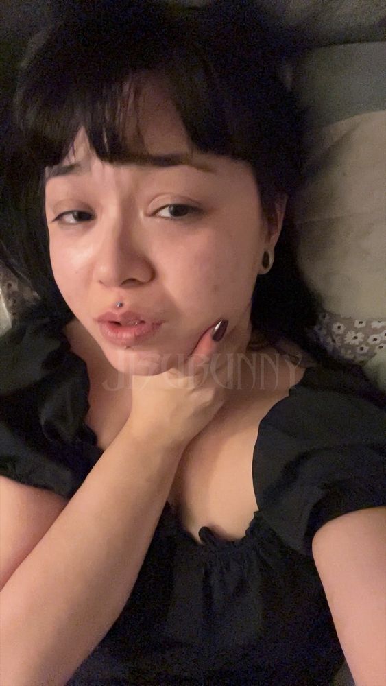 Jisu laying back in a black bar maid dress, with their hand wrapped around their throat and jaw, looking at the camera with pleading eyes