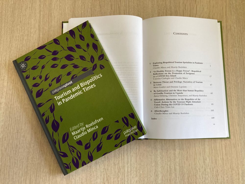A book with a green purple cover that has 'Tourism and Biopolitics in Pandemic Times' written on it, along with the names of the editors 'Maartje Roelofsen and Claudio Minca.. The book lies on top of a table. Underneath the book lies another copy of the book, which is open and shows the 'contents' page of the book.