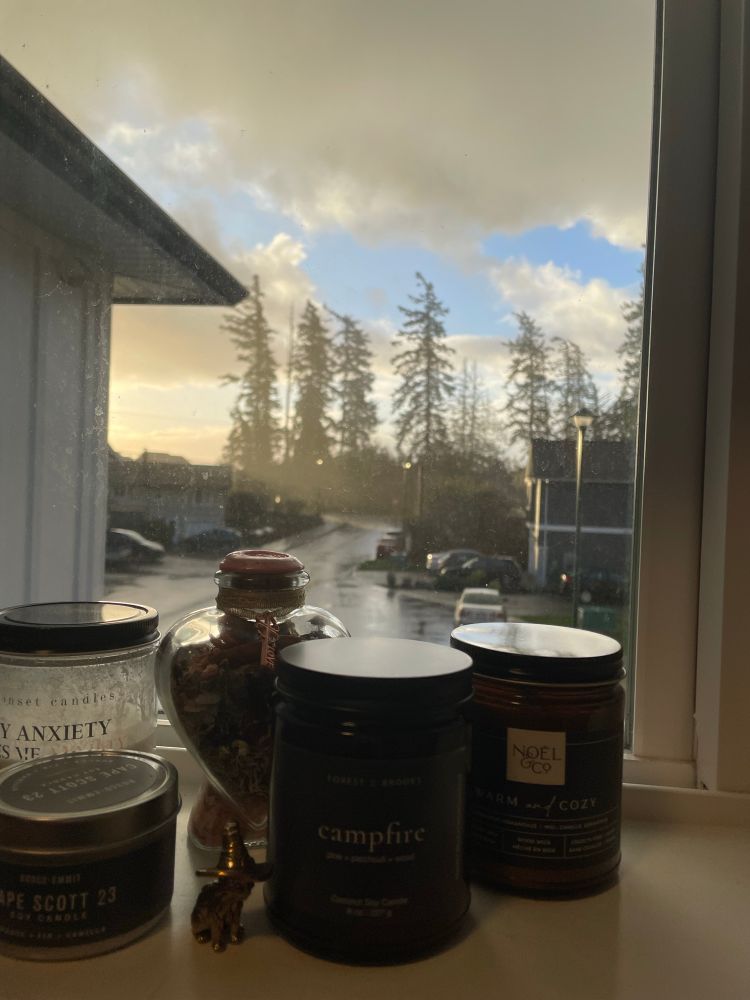 A view, through a window, of a sunny morning and pouring rain. There are various candles on the windowsill in the foreground.
