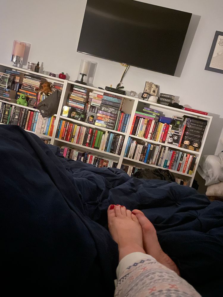 Partial shot of legs and feet, stretched out over a dark blue blanket, and a wall-mounted tv over bookcases.