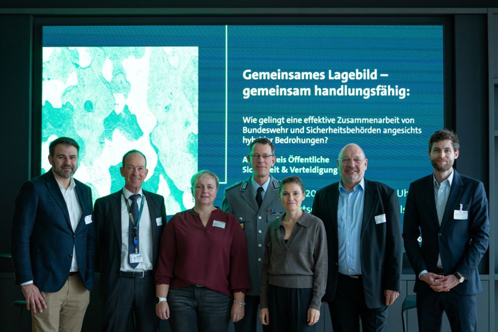 Sieben Personen stehen nebeneinander vor einem großen Bildschirm und posieren für ein Gruppenfoto. Drei Männer in Anzügen, eine Frau in bordeauxroter Bluse, eine Frau mit kurzem Haar in grauem Pullover sowie zwei Männer in Bundeswehr-Uniform. Im Hintergrund ist der Titel der Veranstaltung „Gemeinsames Lagebild – gemeinsam handlungsfähig“ zu lesen.