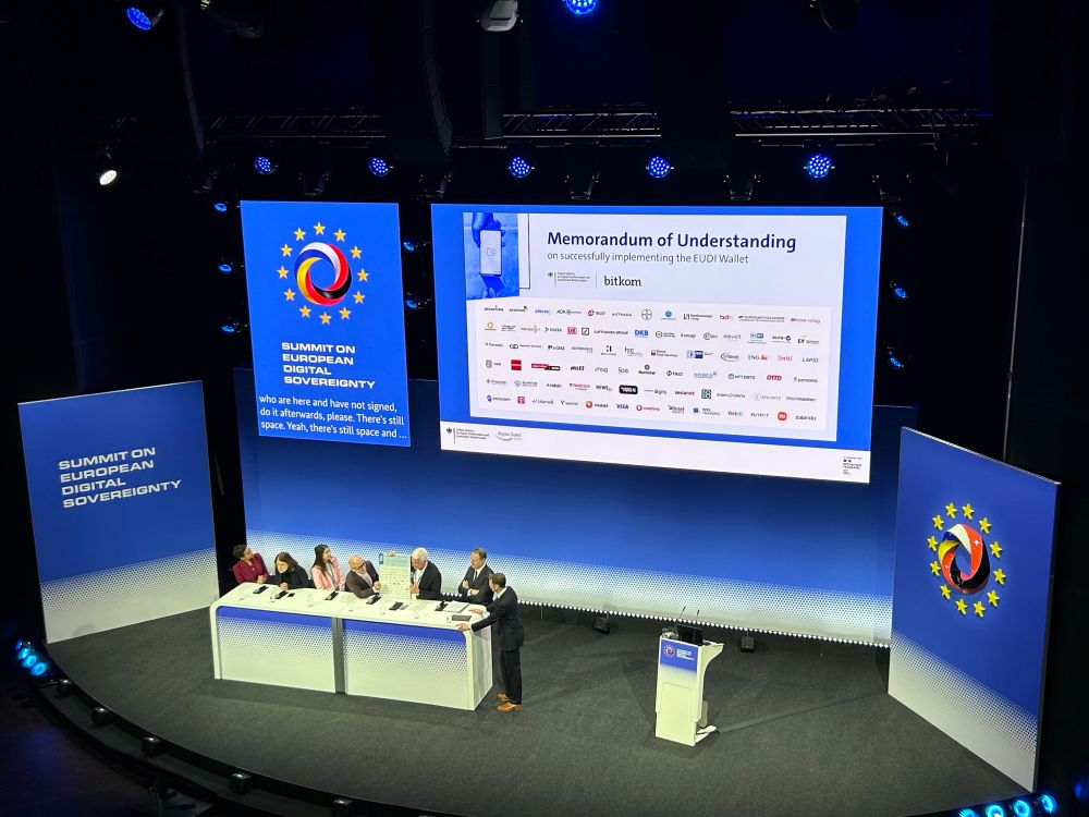 Man sieht die Bühne des europäischen Gipfels zur digitalen Souveränität, in blauem Bühnendesign mit großer Leinwand in der Mitte. Auf der Leinwand ist eine Folie angezeigt, die das „Memorandum of Understanding“ von Bitkom und BMDS zeigt, inklusive diverser Logos unterzeichnender Unternehmen. Vor der Leinwand stehen der Moderator und sechs Bühnengäste, einer davon ist Bitkom-Präsident Dr. Ralf Wintergerst.