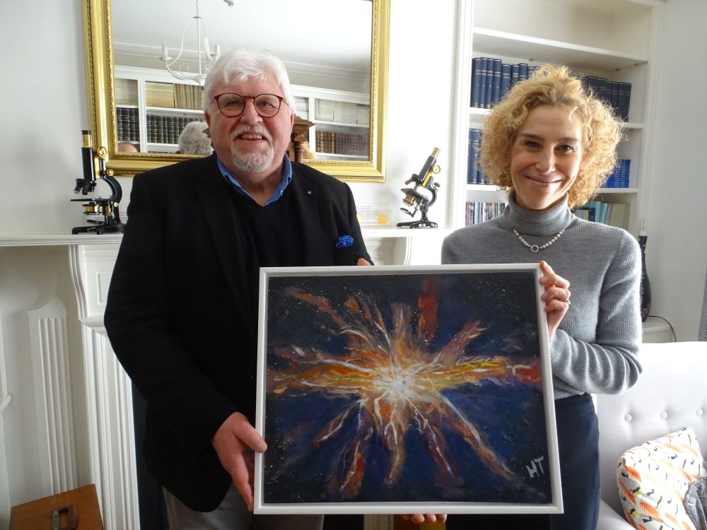 Huw Thomas (left, wearing dark jacket and glasses) holds his painting of a supernova alongside RMS Chief Executive Sali Davis (wearing grey jumper) in the RMS library. A large mirror and mantlepiece with two old microscopes is visible in the background.