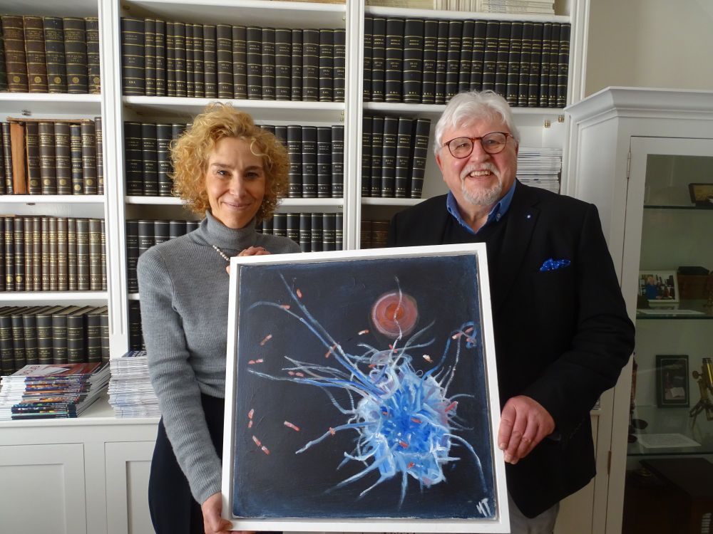 Huw Thomas (right), wearing dark jacket and glasses, holds his painting of a macrophage alongside RMS Chief Executive Sali Davis (wearing grey jumper) in the RMS library. Bookshelves filled with scientific journals are visible in the background.