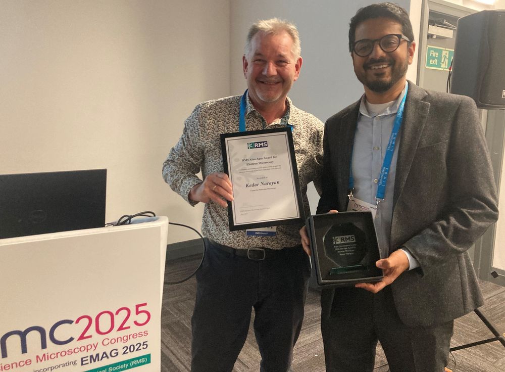 Kedar Narayan (right) holds the Alan Agar Award, presented by Paul Verkade (left), who is holding Kedar's certificate. They are both smiling, standing next to a lectern inside a lecture hall.