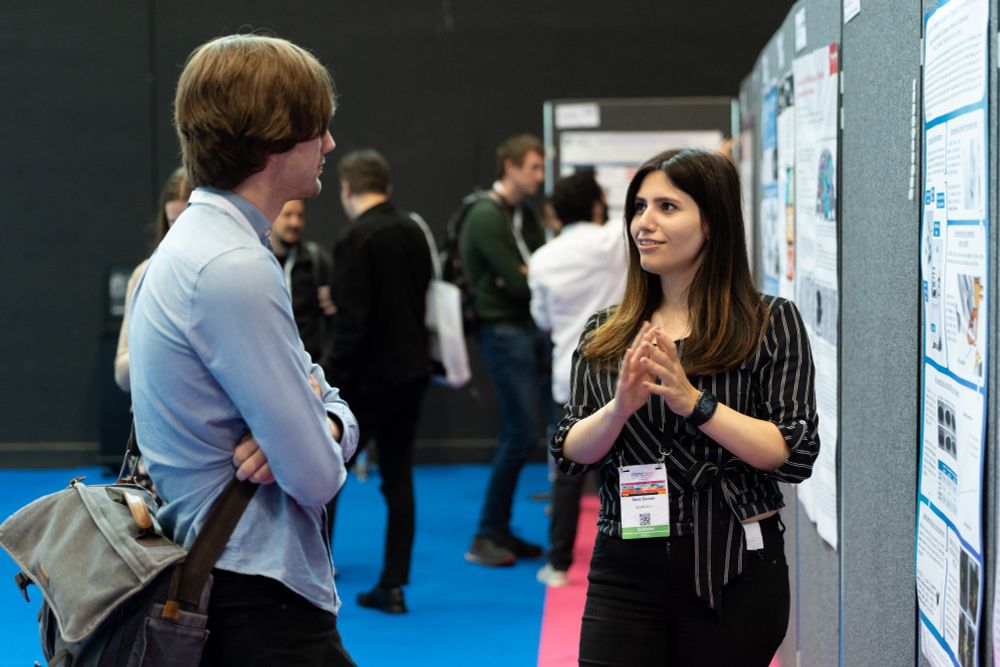 A scientific poster presenter in a black, striped shirt chats with a visitor at mmc2023. A row of posters is visible in the background.