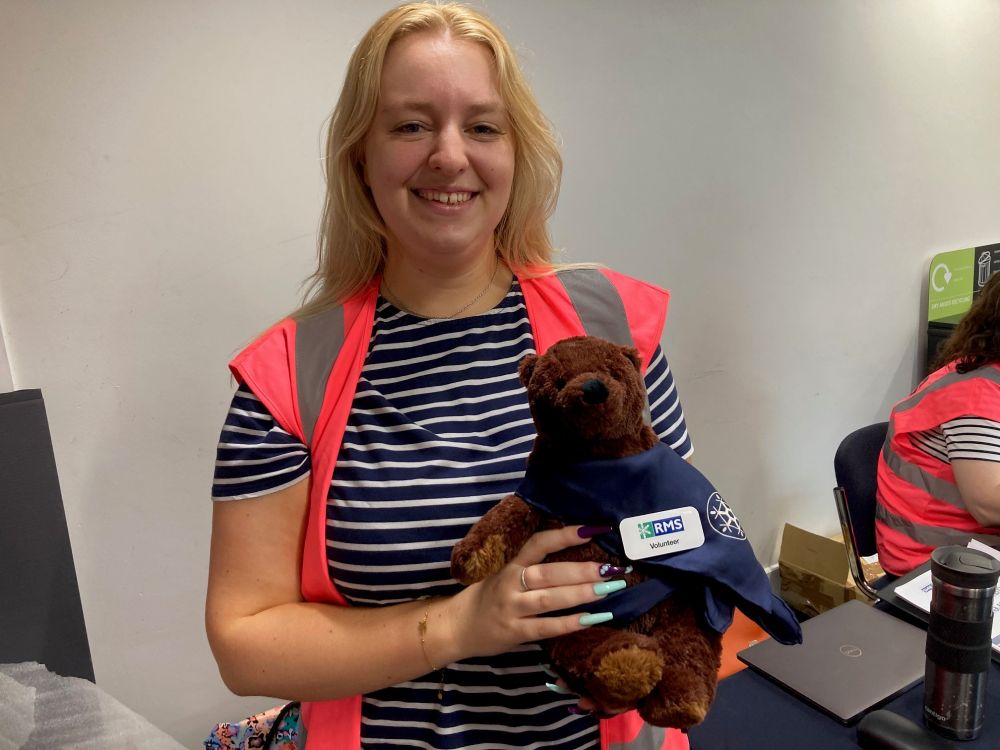 RMS mascot 'Ruggles the Bear' (a small teddy bear) held by Katie from the RMS events team. Ruggles is wearing a blue cape with the RMS snowflake logo on it. Katie is wearing a stripy top and hi-vis vest.