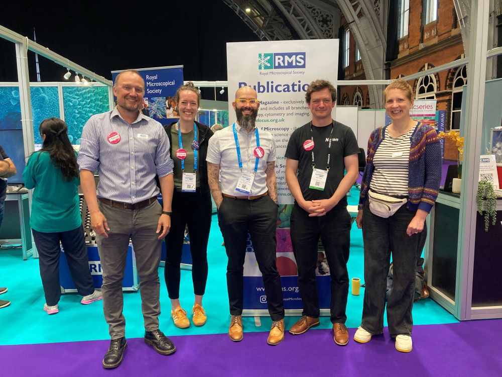 The infocus magazine editorial team pose for a photo in an exhibition hall, next to a large pop-up poster advertising RMS Publications