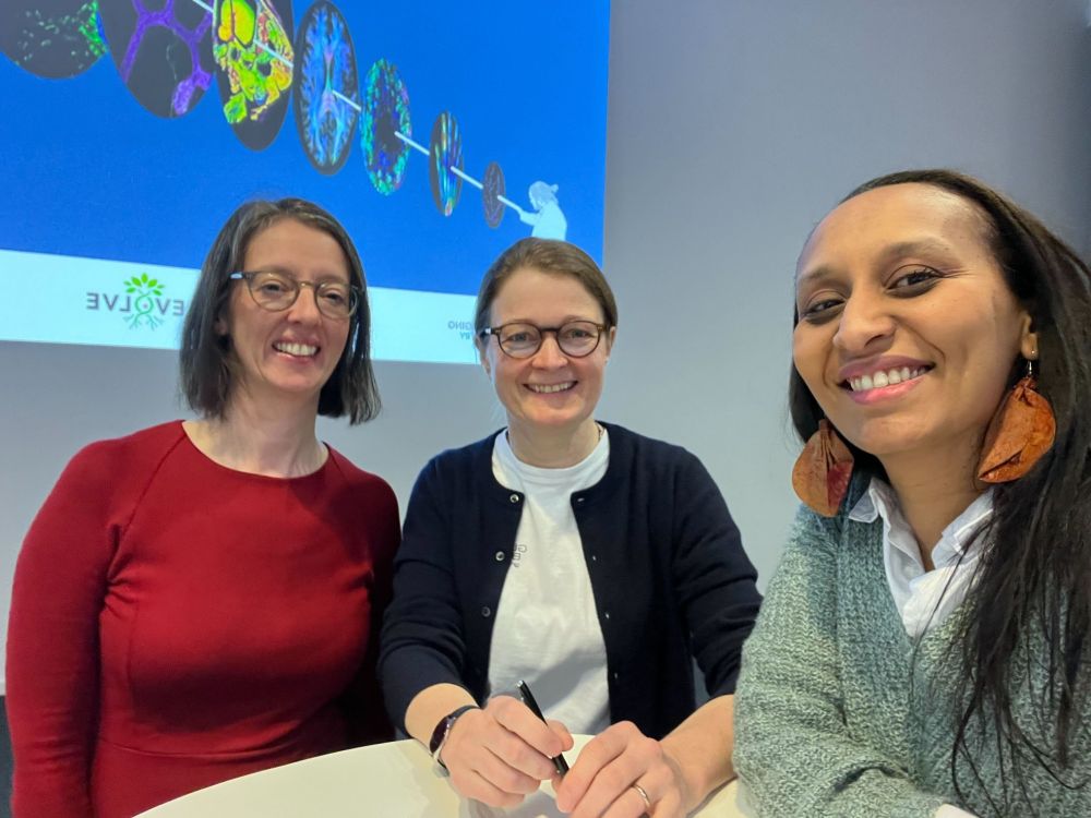 Representatives (three in total, all smiling) of the RMS and Global BioImaging gather around a raised table at the front of a lecture theatre to sign an agreement between the two organisations. A projector screen is visible behind.