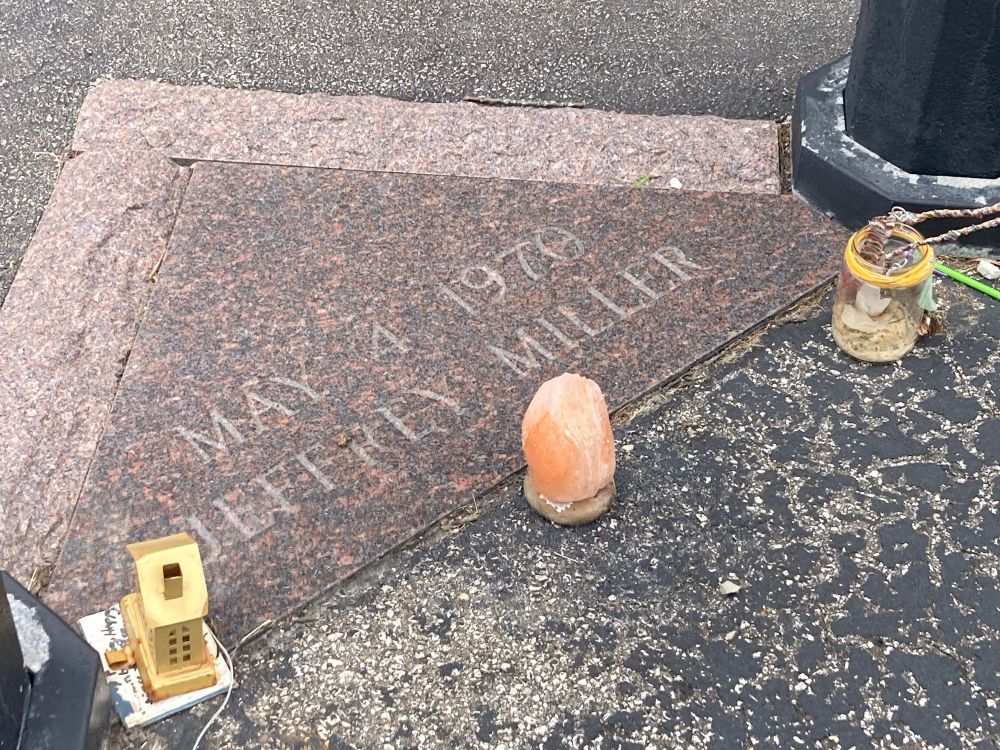 A triangular marker made of pink granite that says. “May 4, 1970 Jeffrey Miller.” There are a couple small, handmade lanterns in the foreground and, near the middle of the picture, a small, pink Himalayan salt chunk. 