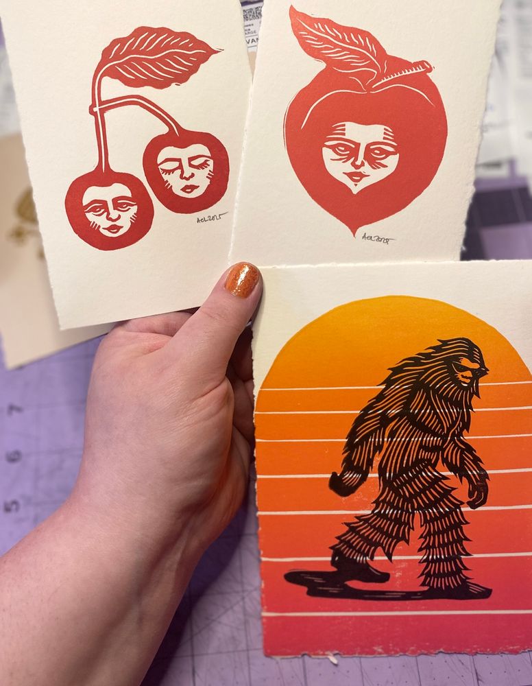 A hand holding three linocut prints; a pair of cherries faces, a peach with a face and a Sasquatch walking in front of a vapor wave-style sunset background 