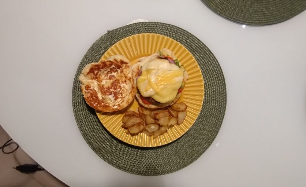 mesa branca com aparador de prato verde e redondo. sobre ele um prato amarelo redondo com um hamburguer com o pão aberto, ao lado dele pedacinhos de batatas assadas.