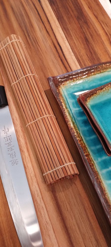 parta da lâmina uma faca yanagi, uma esteira pra fazer sushi e parte de duas peças de louça orientais sobre uma base de corte em madeira 