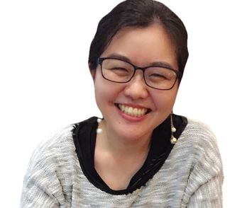 Photo of Emi Tanaka. A smiling woman with dark hair and glasses looks directly at the camera. She has a smile strong enough to make her squint. She is wearing a light-colored sweater over a dark top, with pearl earrings visible.