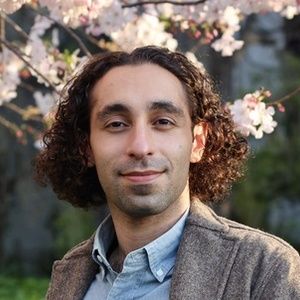 Photo of Nima Hejazi: A man with curly dark hair and a light beard smiles subtly at the camera. He is wearing a light blue collared shirt under a textured brown jacket. Cherry blossoms are visible and slightly out of focus in the background.