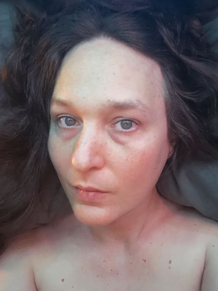 A middle-aged white woman with brown hair taking a selfie in bed under a warm light