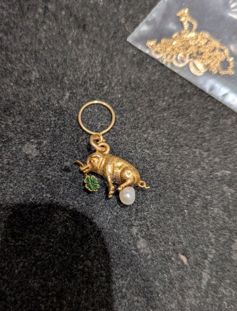 Petit pendentif en or jaune figurant un cochon de profil tenant dans sa gueule un trèfle à quatre feuilles, sous ses pattes arrières une petite perle fine
Au premier coup d'œil je pensais qu'il s'agissait d'un éléphant assis sur une boule, au second on voit bien le cochon qui semble finalement chier une perle (?!)