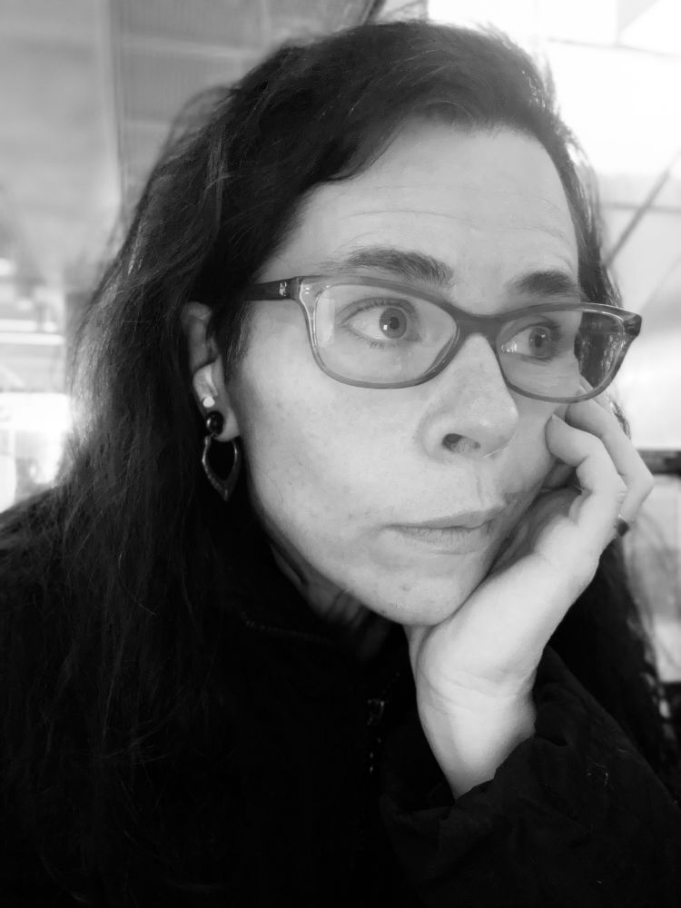 Black and white photo of bemused, dark haired, middle aged woman wearing glasses, staring into the distance. 