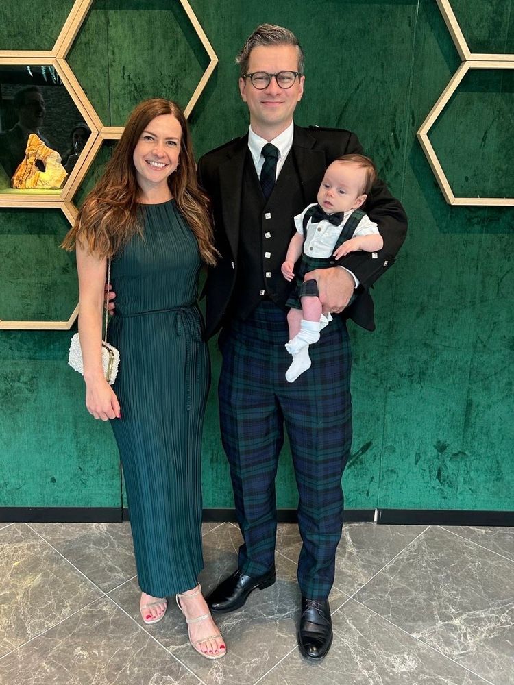 A family stands in front of a green backdrop. A husband, wife, and infant child pose in coordinated formalwear

