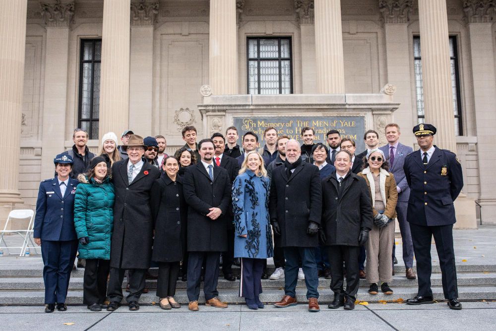 Group of people posing for a photo in front of the Yale Schwarzman Center, with a memorial plaque reading "In Memory of the Men of Yale Who Died in the World War" visible in the background.