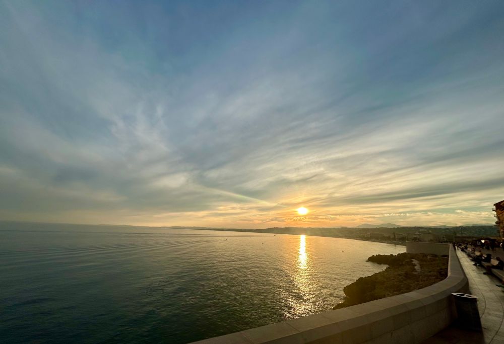 Coucher de soleil sur la mer Méditerranée et le bord de mer avec à droite de la photo la promenade en pierre lisse, la plage et une vue sur une partie de la ville.
