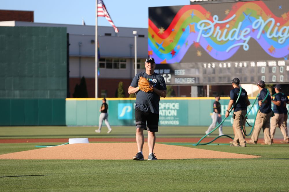 A photo of Commissioner Upthegrove preparing to throw the first pitch. Readying the ball in his glove, with "Pride Night" displayed on the scoreboard behind him. 