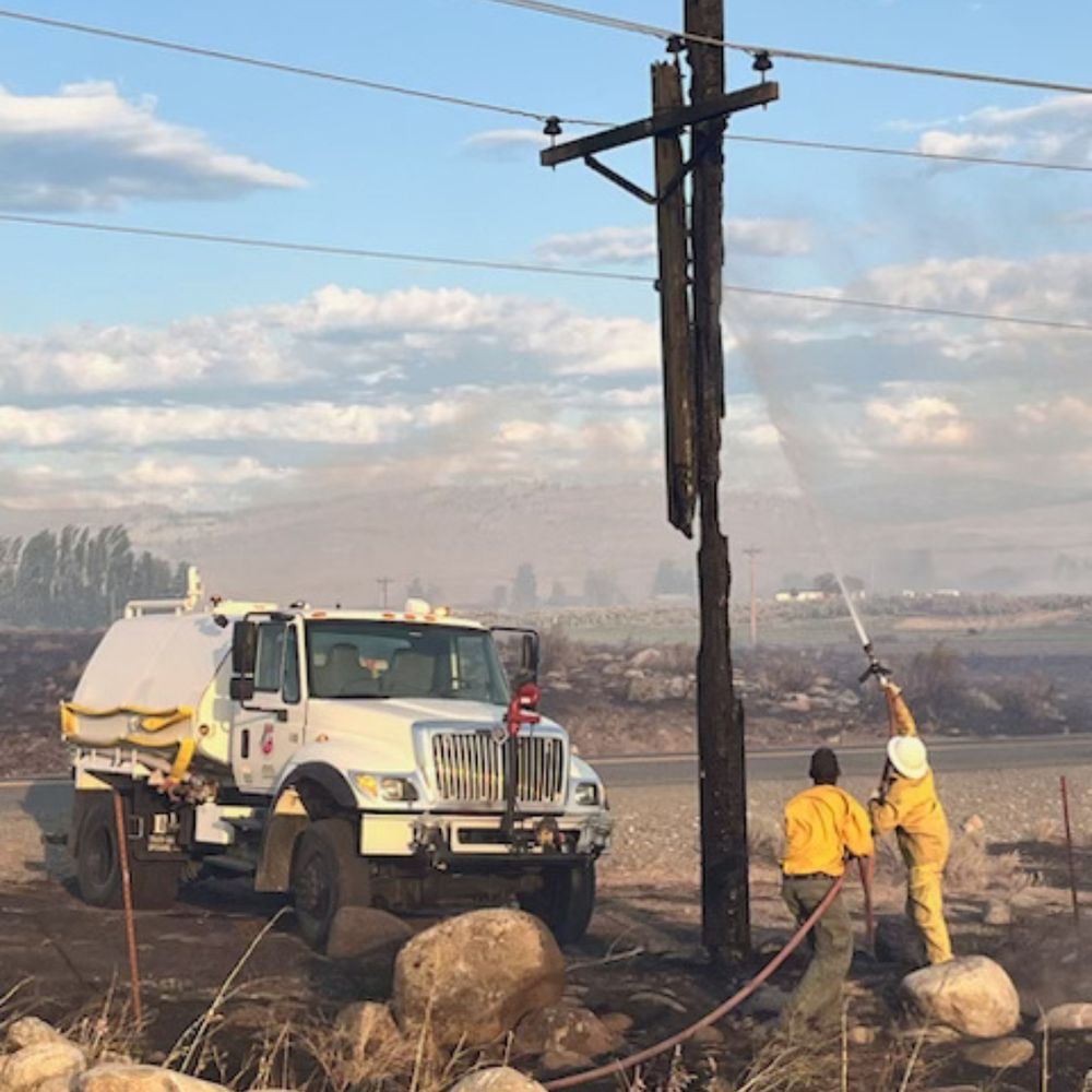A photo of two wildland firefighters working with a hose from a small tank truck to put out a hot spot on a utility pole. Thank you to the Okanogan County PUD for sharing this photo of the efforts happening there. 