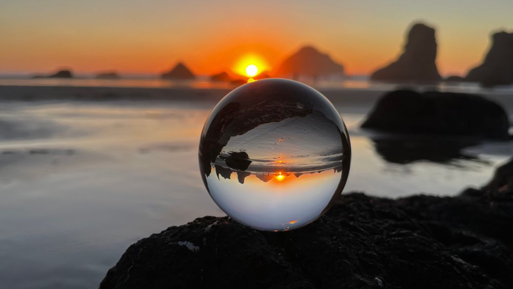 A brilliant orange setting sun and  sea-stacks in the distance, and a clear glass sphere that sits on dark rock in the foreground, reflecting the scene in the distance