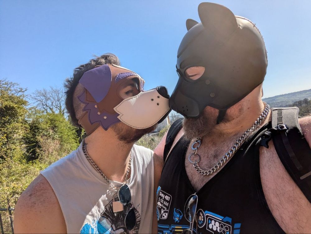 a tender photo of me and pup silas booping our snoots. the sky is bright blue, the sun is shining, and there is an awful lot of love and care in our expressions

we are both pups wearing vests - my hood is open and bright and my vest is white and slutty. their hood is black and enclosed and their vest is black and slutty
