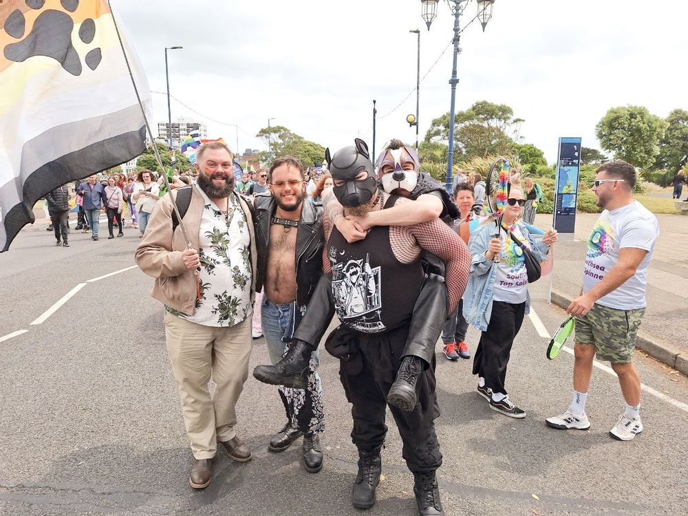 another picture of us in the parade; this time we're posing with our friends. a beautiful bear in a Hawaiian shirt waves a bear pride flag; a sexy cub in leathers and a harness grins for the camera, and I am clinging to Silas's shoulders as they give me a piggyback ride. oink oink